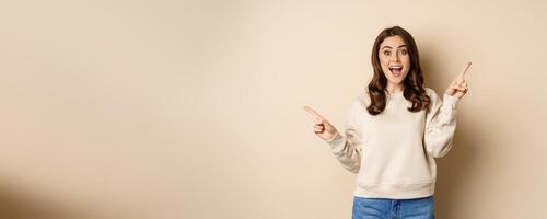 Cheerful caucasian woman showing directions, two ways, pointing sideways at variants, choices for customer, standing over beige background photo