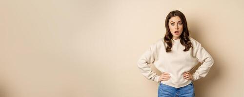 Portrait of shocked brunette woman drop jaw, gasping and staring speechless at camera, beige background photo