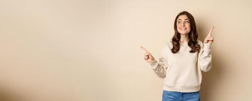 Cheerful caucasian woman showing directions, two ways, pointing sideways at variants, choices for customer, standing over beige background photo