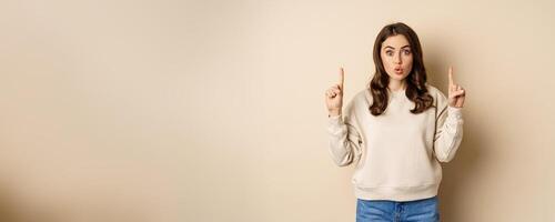 Surprised beautiful girl showing advertisement, pointing fingers on top, store sale or website link up, standing over beige background photo