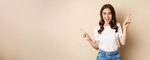 Happy authentic girl smiling, pointing fingers sideways, showing left and right banner, demonstrating promo, standing against beige background photo
