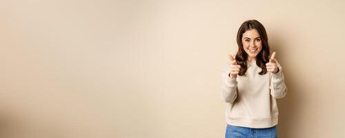 Smiling happy woman choosing you, pointing fingers at camera and laughing, inviting people, standing over beige background photo