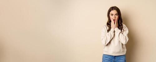 Portrait of shocked brunette woman drop jaw, gasping and staring speechless at camera, beige background photo