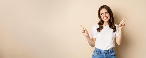 Happy authentic girl smiling, pointing fingers sideways, showing left and right banner, demonstrating promo, standing against beige background photo