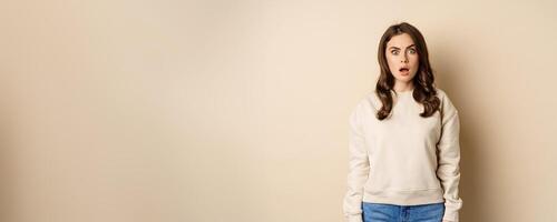 Portrait of shocked brunette woman drop jaw, gasping and staring speechless at camera, beige background photo