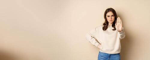 Stop. Serious and confident woman showing extended arm palm, prohibit, forbid smth, blocking something, standing over beige background photo
