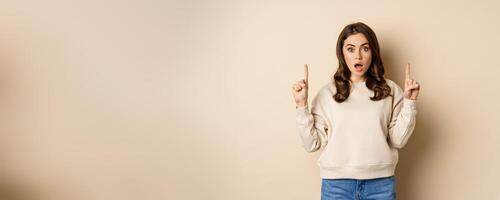 Surprised beautiful girl showing advertisement, pointing fingers on top, store sale or website link up, standing over beige background photo