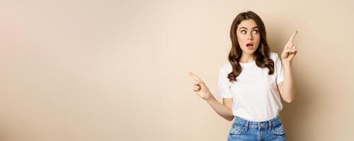 Happy authentic girl smiling, pointing fingers sideways, showing left and right banner, demonstrating promo, standing against beige background photo
