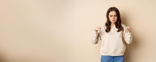 Sad and disappointed girl pointing, looking down with upset grimace moody face, standing over beige background photo