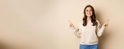 Cheerful caucasian woman showing directions, two ways, pointing sideways at variants, choices for customer, standing over beige background photo