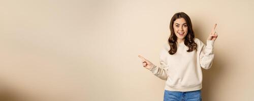 Cheerful caucasian woman showing directions, two ways, pointing sideways at variants, choices for customer, standing over beige background photo