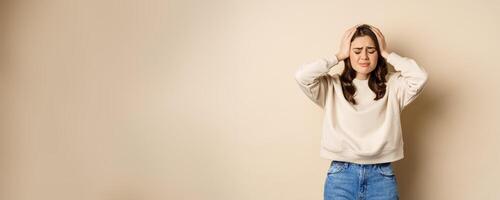 Frustrated and troubled woman facing disaster, holding hands on head in panic, looking anxious, standing over beige background photo