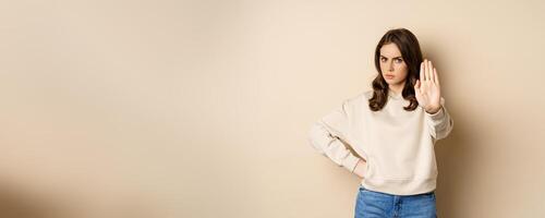 Stop. Serious and confident woman showing extended arm palm, prohibit, forbid smth, blocking something, standing over beige background photo