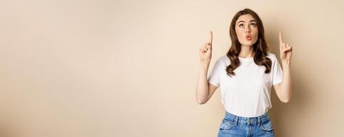 Surprised brunette girl showing promo, pointing and looking up with amazed face expression, standing over beige background photo