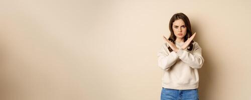 Woman showing stop, cross sign gesture and frowning angry, dislike and disapprove, saying no, rejecting and prohibiting, standing over beige background photo