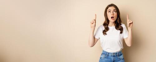 Surprised brunette girl showing promo, pointing and looking up with amazed face expression, standing over beige background photo