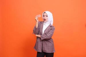 Young indonesia woman in hijab suit surprised with arms crossed while showing isolated plastic debit credit card on orange background. customer satisfied with service for financial, mobile transaction photo