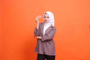Young Asian girl in hijab suit frowning with arms crossed while showing isolated plastic debit credit card on orange background. customer satisfied with service for financial , mobile transaction photo