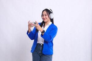 beautiful adult office woman wearing blue formal suit standing on right side smiling playing game on smartphone mobile phone and headphone isolated white background. for technology, entertainment photo