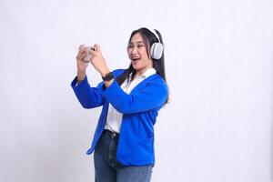 beautiful adult office woman wearing blue formal suit standing on right side happily playing game on smartphone mobile phone and headphone isolated white background. for technology,entertainment photo