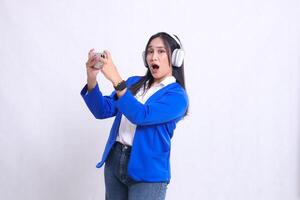 beautiful adult office girl wearing blue formal suit standing right sideways surprised to camera both hands playing game on smartphone mobile phone and headphone isolated white background. technology photo