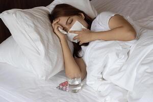 A woman lying in bed feeling ill, with a glass of water, tissues, and medicine beside her photo