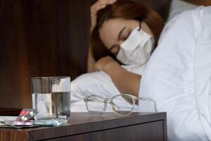An Asian young woman wearing a mask, appears dizzy. Selective focus on the pills and glass. photo