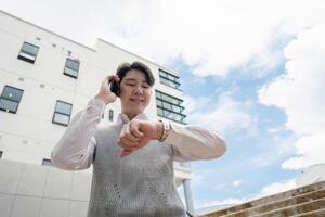 Young Professional Checking Time While Talking on Phone Outside Modern Office Building on a Sunny Day photo