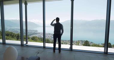 A man near a panoramic window, enjoying view of a serene lake and majestic mountains video