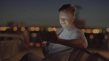 A man sits on a city park bench at night, using a tablet with bokeh lights in the background video