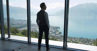 Man Staring at Scenic Mountain Lake View through Large Window in Modern Building video