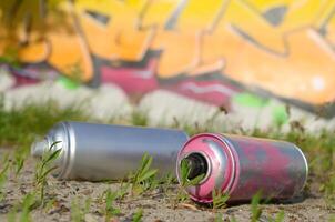 Several used spray cans with paint and caps for spraying paint under pressure on grass near the painted wall in colored graffiti photo