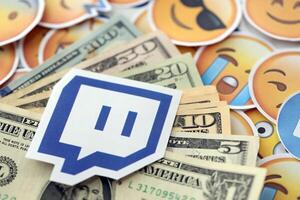 KYIV, UKRAINE - APRIL 20, 2024 Twitch logo and many usd money bills. Symbol of famous social network platform and dollar currency banknotes on lot of emoji reactions photo