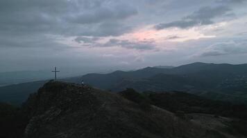 Aerial view caucasian woman in white stand on viewpoint mountain hilltop with giant standing cross on dramatic sunset and cloudy evening in nature. Believer search for purpose hope and pray conceptual video