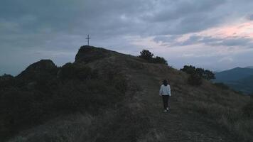 Aerial view caucasian woman in white walk uphill to mountain hilltop with giant standing cross on dramatic sunset and cloudy evening in nature. Believer search for purpose hope and pray conceptual video