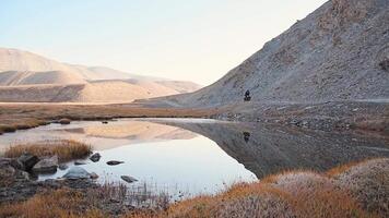 Slow motion zoom in front view adventurous traveler cycle bicycle touring solo on gravel road in remote high Pamir mountains early morning by river. Pamir highway Bartang valley video
