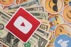 KYIV, UKRAINE - APRIL 20, 2024 YouTube logo and many usd money bills. Symbol of famous social network platform and dollar currency banknotes on lot of emoji reactions photo