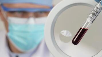 Doctor holds a test tube with a blood sample. Scientist in a medical mask sits in front of a microscope. video