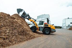 industrial wheel loader with raised bucket at a biomass power plant, handling wood chips against a backdrop of storage silos and blue sky, showcasing renewable energy resources photo