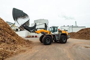 industrial wheel loader with raised bucket at a biomass power plant, handling wood chips against a backdrop of storage silos and blue sky, showcasing renewable energy resources photo