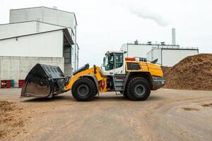 industrial wheel loader with raised bucket at a biomass power plant, handling wood chips against a backdrop of storage silos and blue sky, showcasing renewable energy resources photo