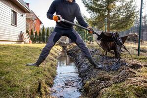 Determined man shoveling mud from a trench in his backyard on a sunny day, with a wheelbarrow and rural home in the background photo
