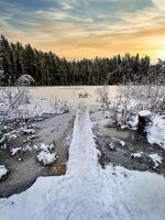 A winter wonderland scene with a frozen lake, a wooden bridge and a stunning sunset in the background The colors of the sky are a mix of orange, pink and purple hues creating a serene atmosphere photo