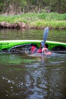adventurous kayaker in a bright green kayak practicing the eskimo roll technique in a calm river surrounded by lush vegetation during an overcast day photo