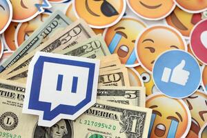 KYIV, UKRAINE - APRIL 20, 2024 Twitch logo and many usd money bills. Symbol of famous social network platform and dollar currency banknotes on lot of emoji reactions photo