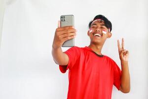 A young Asian man with a happy successful expression while holding his phone and wearing red top, flag headband and cape isolated by white background. Indonesia's independence day concept. photo