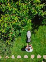 Caucasian gardener mowing backyard garden grass using Cordless electric grass mower. Aerial View. photo