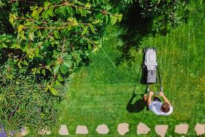 Caucasian gardener mowing backyard garden grass using Cordless electric grass mower. Aerial View. photo