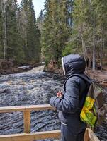 woman traveler with backpack exploring beautiful pine forest river. girl on hiking trip looking at waterfall in coniferous forest in mountains. concept of spring or autumn travel, nature exploration photo