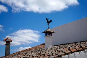 A weather vane shaped like a rooster sits atop a chimney against a cloudy sky, adding a rustic charm to the rooftop scenery. photo